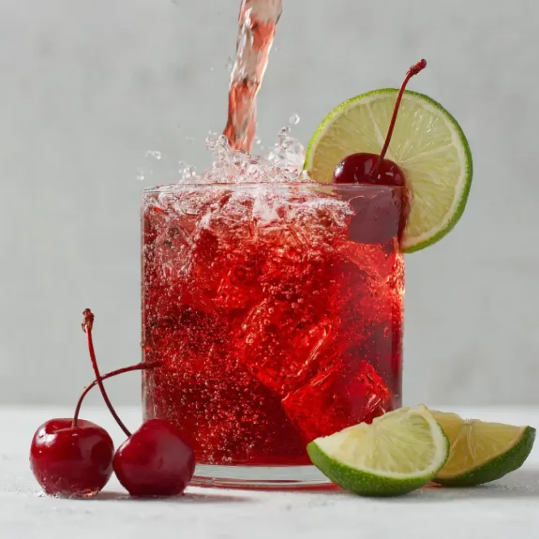Vibrant le cocktail shirley being poured into a glass with lime and cherries.