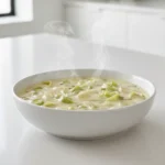 Fondue de Poireaux Express servie dans un bol blanc sur un plan de travail immaculé, dans une cuisine moderne blanche, avec de la vapeur s’échappant du plat.